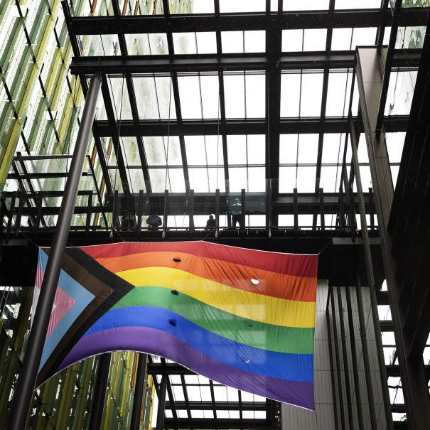 An image of the new Progress Pride flag flying in the breezeway of one of Amazon's offices at its Seattle headquarters. The flag features the rainbow on the right and a triangle with light pink and blue and black and brown on the left.