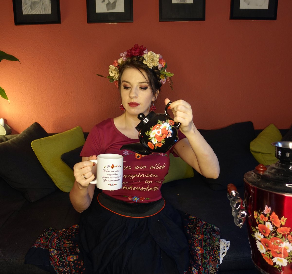 Marina gießt Tee ein in ihre "Wenn wir alles anzünden, sparen wir uns viele Zwischenschritte"-Tasse. Sie hat einen Blumenkranz auf dem Kopf und ist fancy as hell.