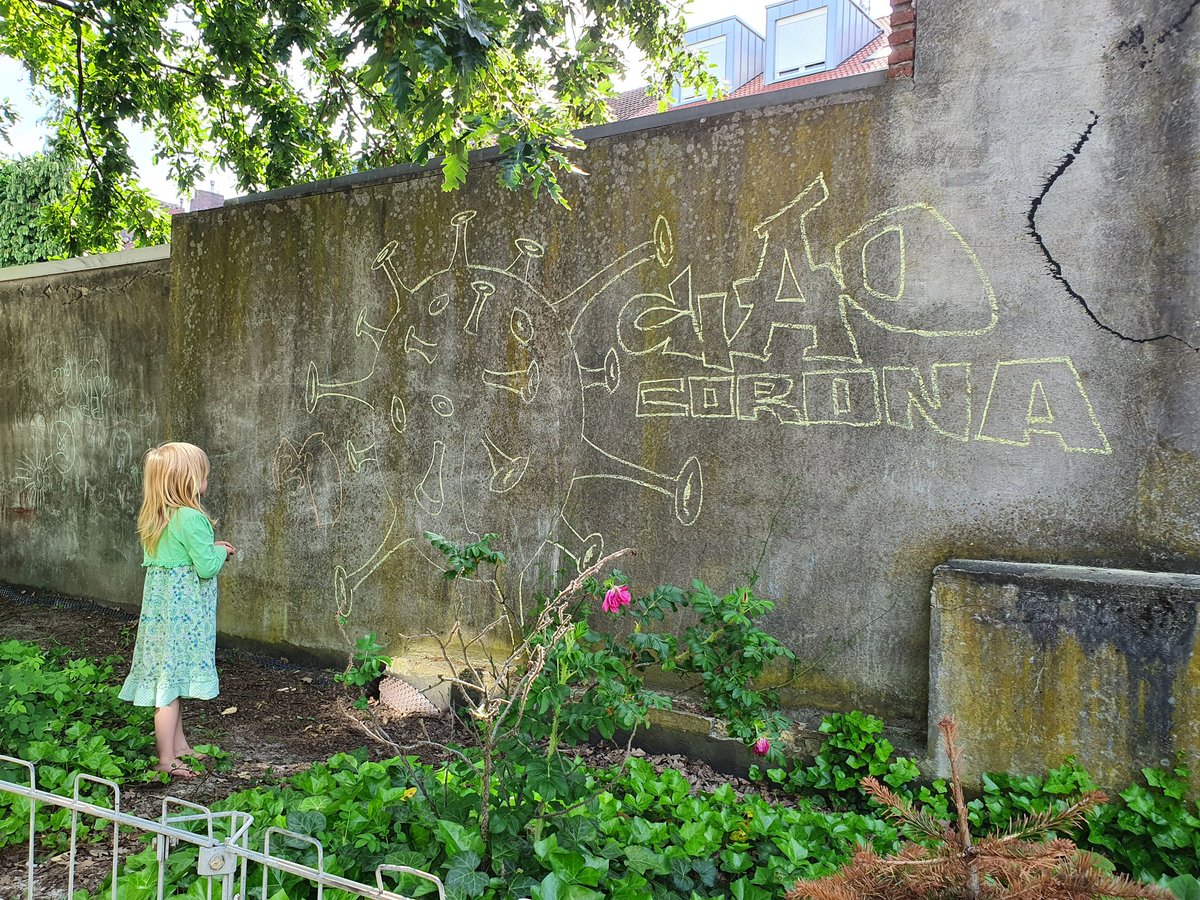 Foto. Ein kleines Mädchen schaut auf eine Mauer. Darauf ist mit Kreide ein großes Corona-Virus gemalt. Dabei steht "Ciao Corona"