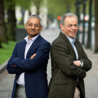 Two men stand with their arms crossed, with trees in the background.