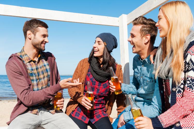 Four happy friends talking to each other and smiling while sitting on the beach and drinking beer.