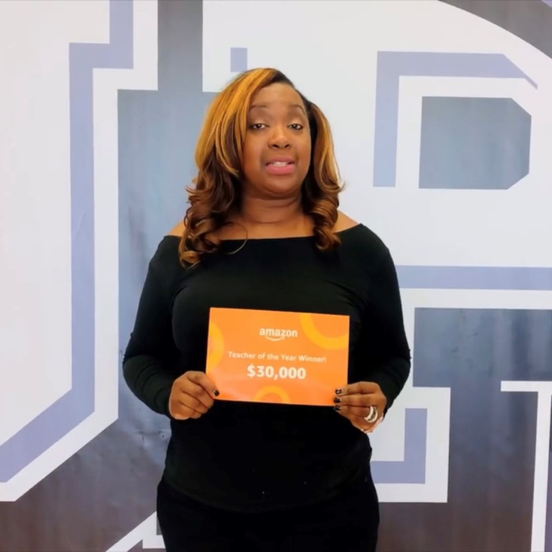 Melissa Collins wears a black dress and holds up an orange certificate that reads, "Amazon Teacher of the Year Winner $30,000." 