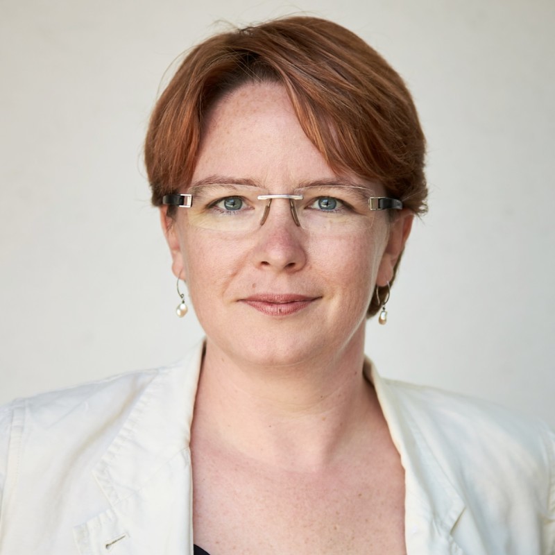 A woman smiles for a headshot photo in front of a white background while wearing a white blazer and glasses.