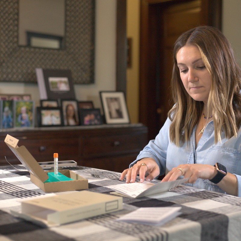 An image of a woman sitting at her kitchen counter with an at-home COVID test in front of her. She is reading directions that came with the test.