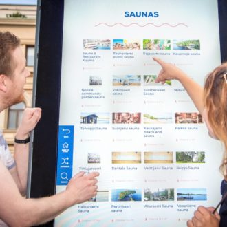 A woman and a man point at pictures on a large outdoor screen.