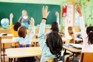 Young school children with hands raised in a classroom setting seen from behind.