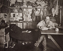 Man sitting cross-legged at table, pipe in hand, apparently writing. Much clutter of clothing, books and equipment is in the background.