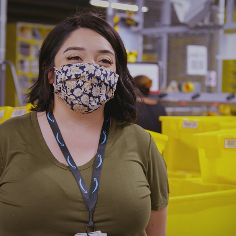 A woman wearing a t-shirt, lanyard, and face mask stands in an Amazon fulfillment center.