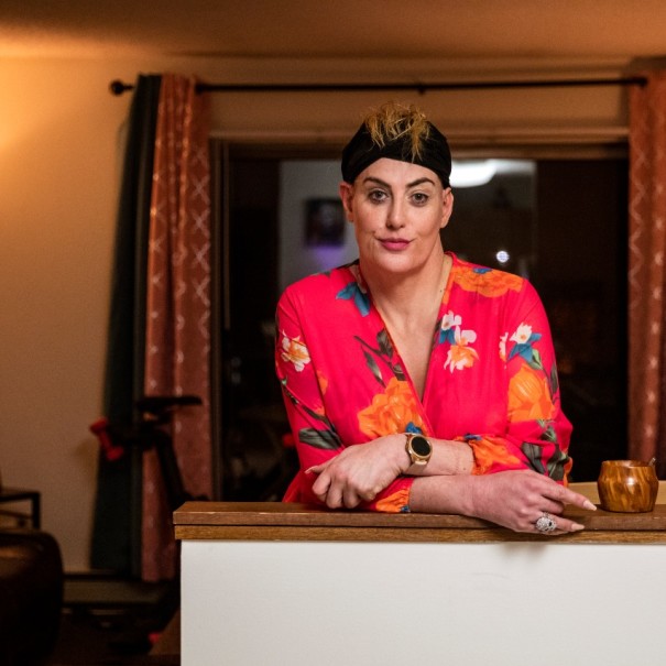 An image of a woman softly smiling for a photo while standing at the kitchen counter in her apartment. She is wearing a black headband and a pink floral robe.