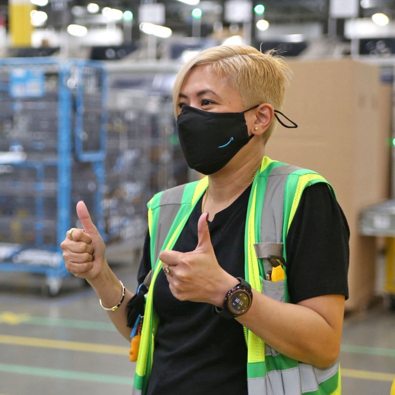 A woman wearing a face mask and a safety vest gives two thumbs up to someone off-camera. She is standing in an Amazon fulfillment center.