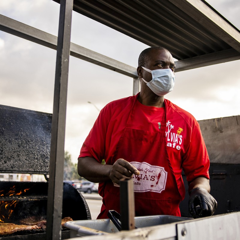 A man stands in front of an outdoor smoker. He wears a mask and holds products to prepare meals for customers. 