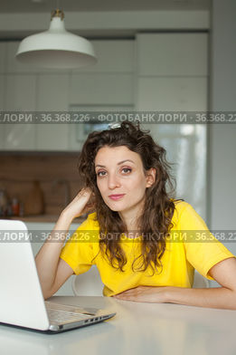 Young woman with curly hair and yellow shirt is working from home using her laptop at the kitchen table in her apartment, remote work, freelance