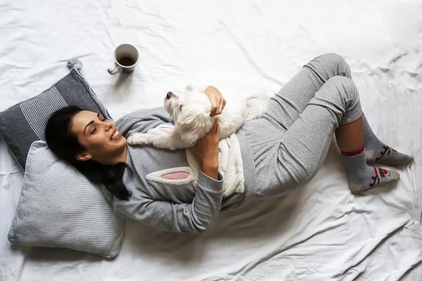 Woman relaxing on a bed cuddling a white dog with cup of coffee next to her.