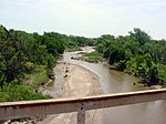 A small, muddy river, as seen from a bridge, meanders between tree-lined banks.