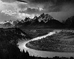 A river winds across a plain at the foot of jagged snow-covered mountains.