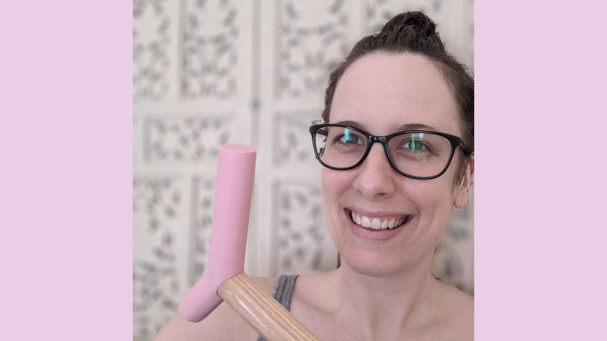 Graphic with a soft pink background featuring a photo of Brianne, a white woman in her mid-30s wearing black framed glasses and dark brown hair in a messy bun, smiles at the camera. She’s wearing a grey tank top, holding the pink epoxy handle of her blonde wood cane up beside her face and sitting in front of a blurred off-white decorative screen. The photo has been cropped close around Brianne’s face, shoulders and cane handle.