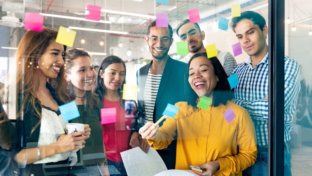 A group of diverse colleagues brainstorming ideas in front of a glass wall using post-it notes.