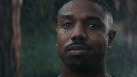 Michael B. Jordan has droplets of water on his face, and the background looks to be outdoors, with driving rain. His eyes have a blue hue to them. 