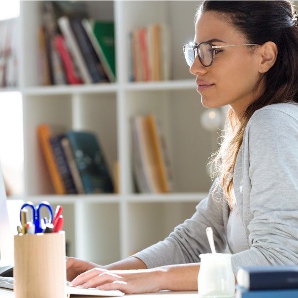 A woman wearing glasses sits at a computer, she is looking at the monitor and her hands are on the keyboard. 