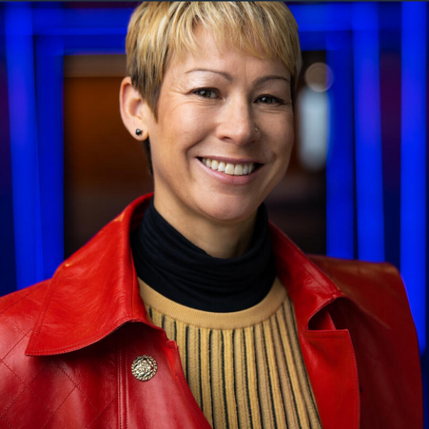 An image of a woman smiling for a headshot in a hallway of blue lights. She is wearing a red jacket and yellow sweater. 