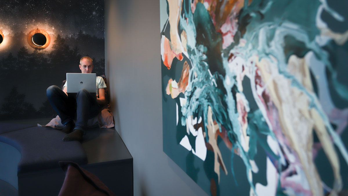 Young man wearing a white T-shirt sitting with his feet up on a large bean bag with a laptop on his lap. Colorful abstract painting on the wall.