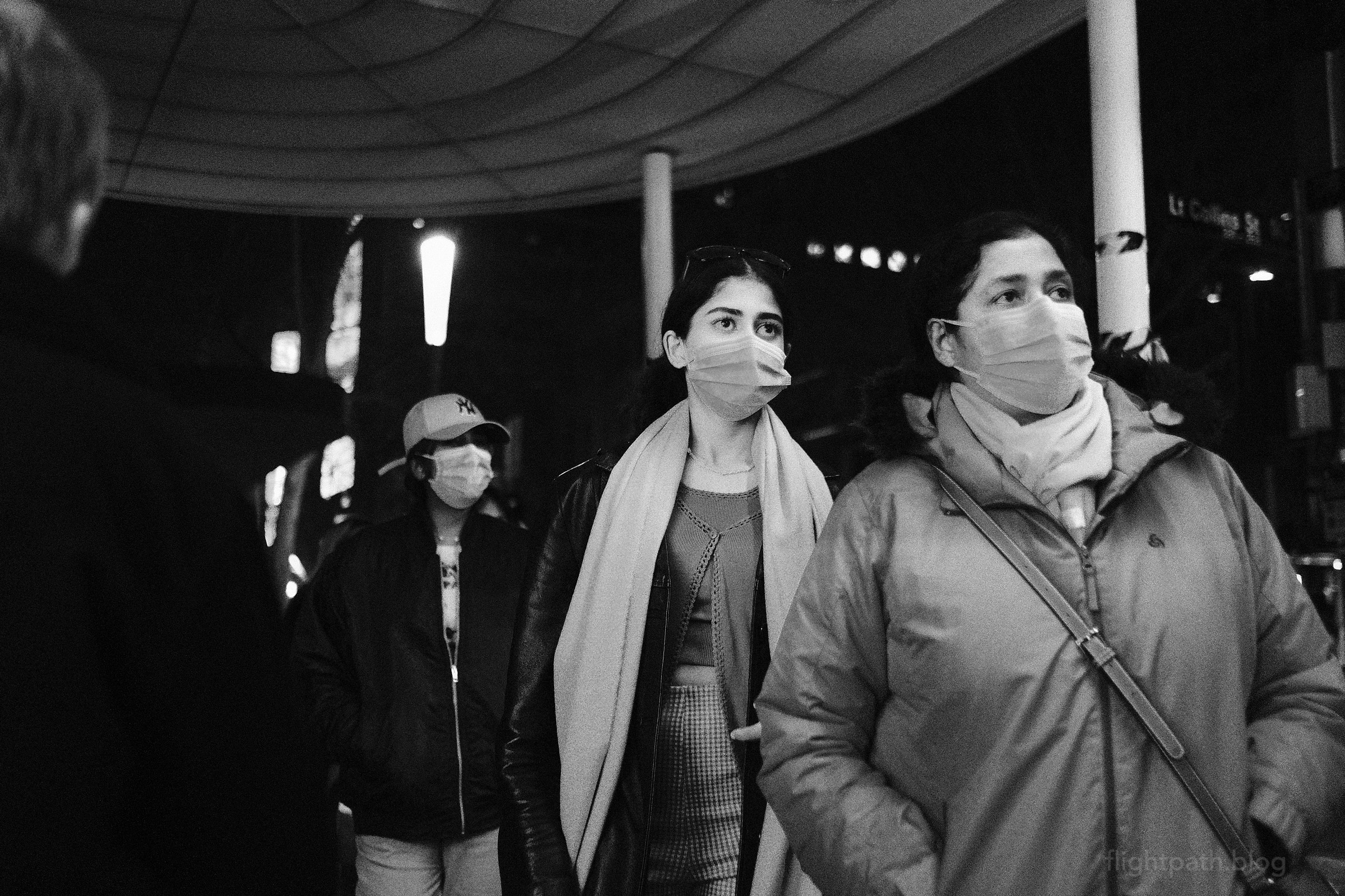 Three people wearing disposable masks are all facing the same direction. It is night. A street sign, barely visible, labels Little Collins Street.