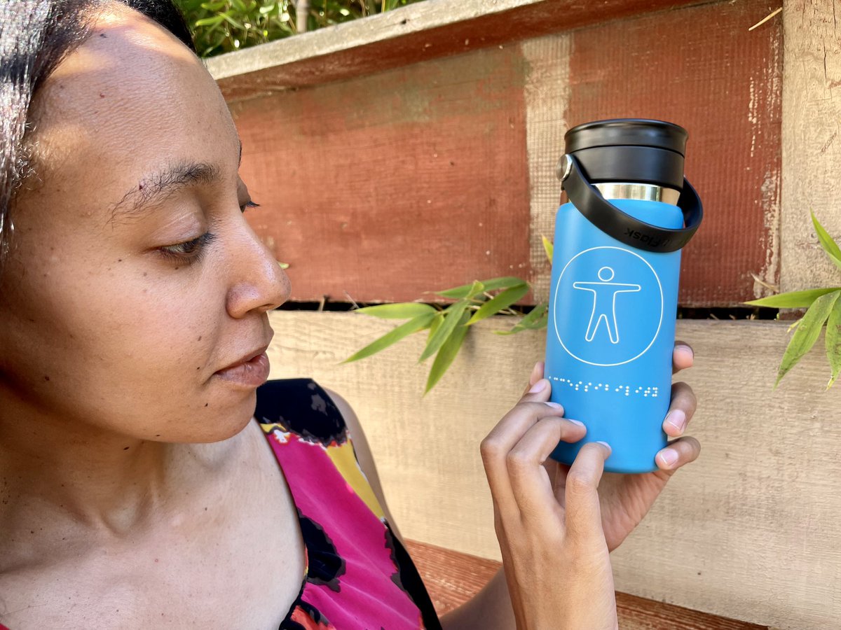 I’m holding up and reading the word on the thermos. It’s all blue with white raised markings. There is a figure of a person in a circle, the universal symbol for accessibility. Below that are braille letters for the word: accessibility.