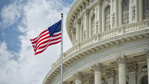 The United States Capitol, against a bright blue sky with fluffy clouds. A U.S. flag flaps in the wind in the foreground. 