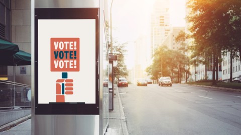 A metal bus station on a city street, with a illustrated poster on it tyat says "vote! vote! vote!" 