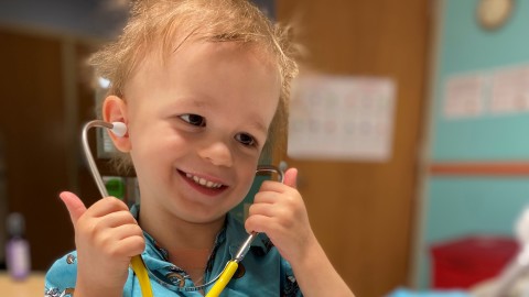 An image of a young boy smiling while hold a stethoscope to his ears.