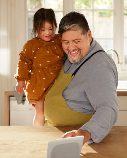 The father holds his toddler daughter in the kitchen and touches the Nest Hub display as they prepare a recipe.