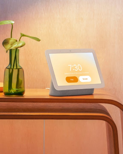 Nest Hub is on top of a bedroom counter. The screen displays information that is backlit by a gentle orange glow.