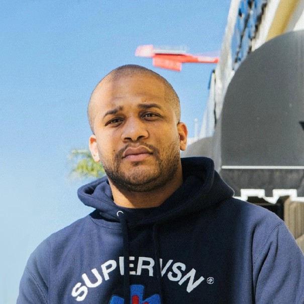 An image of a man standing for a photo outside. There is a palm tree and a street view behind him.