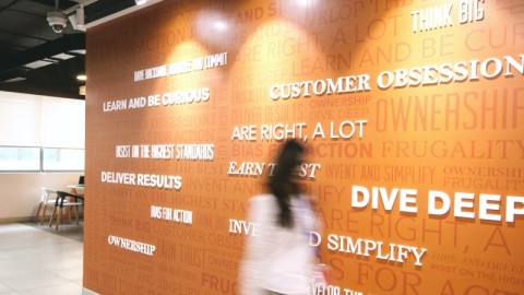 Amazon's Leadership Principles featured on a wall in an Amazon office building. A woman is walking in front of them.