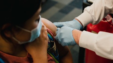 A woman wearing a mask holds up her sleeve as she receives the COVID-19 vaccine from a nurse wearing a white jacket and blue gloves.