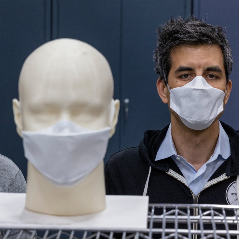 Two men wearing face masks stand behind a mask prototype.