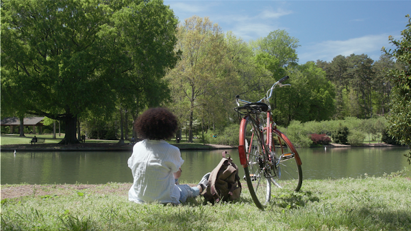 Girl with her bike sitting in the grass overlooking a tree-lined pond on a sunny day