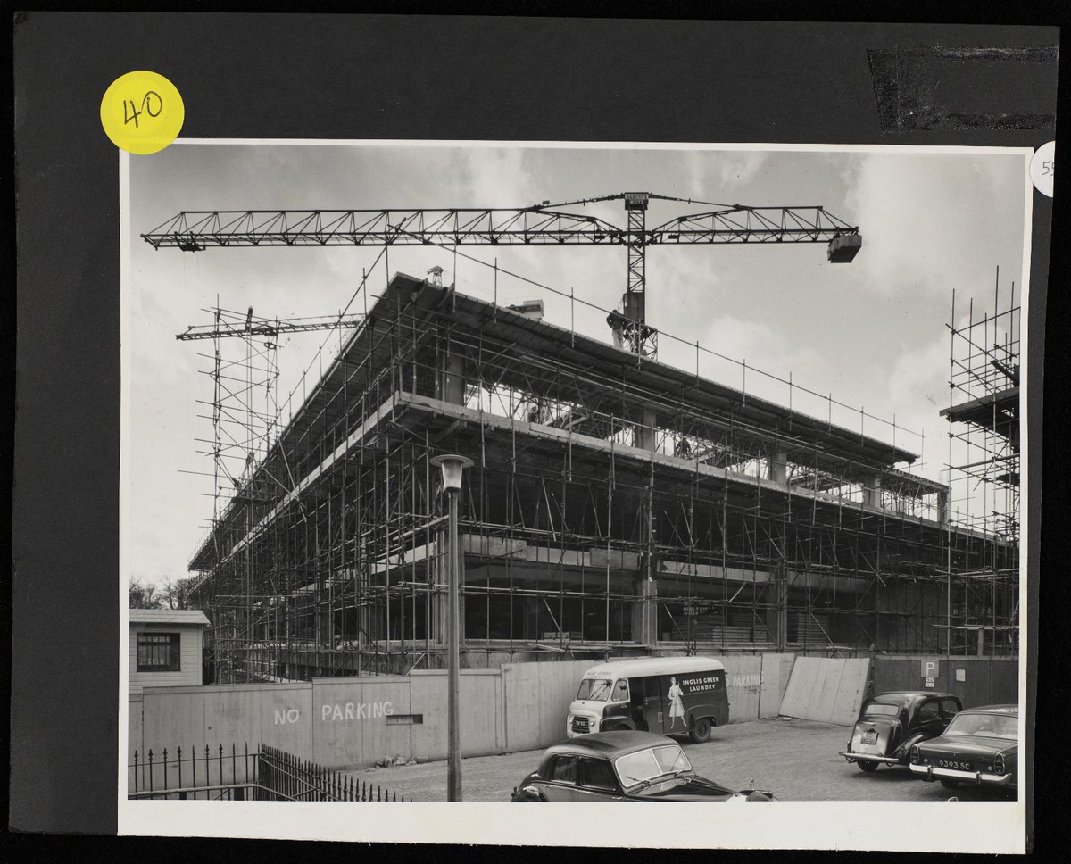 Black and white photograph of construction of Main Library from 1965-1967. In this photo the shell of the building is being built and is surrounded by scaffolding and cranes. In the foreground are several vehicles from the 1960s.