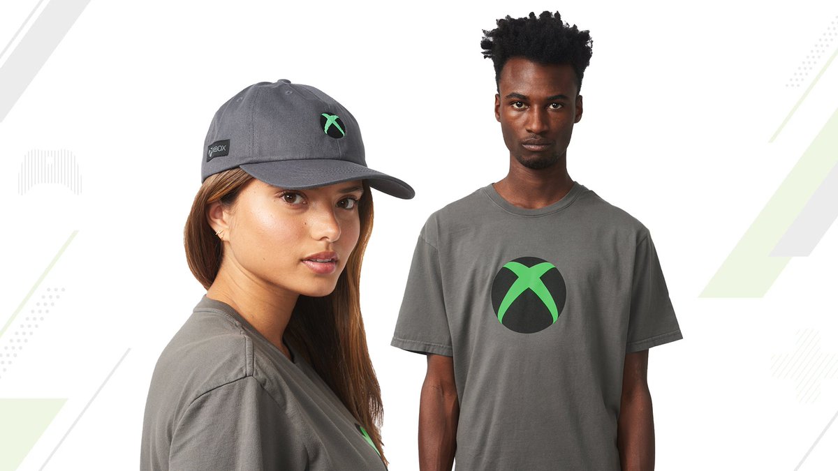 A female model stands in the foreground looking to camera wearing at the camera wearing a grey baseball cap with a green-and-black Xbox nexus logo on it, reading Xbox on the side. A male model stands in the background wearing a t-shirt in the same grey with the same green-and-black Xbox nexus logo printed on the front.