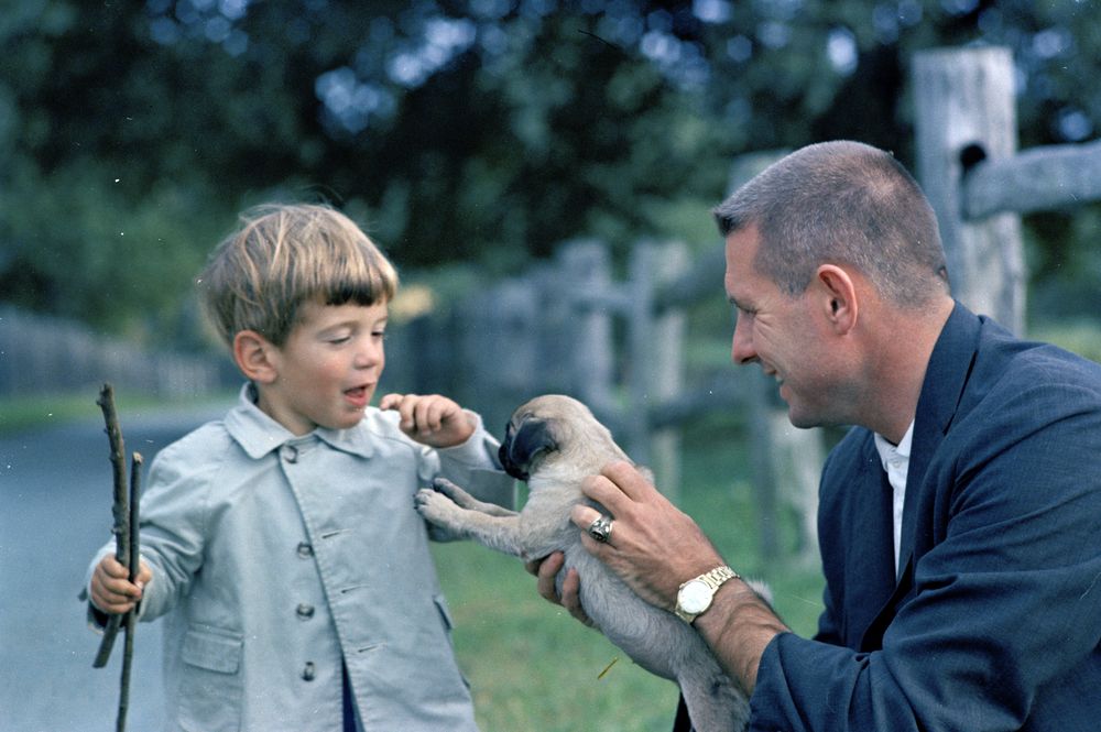 Secret Service agent Bob Foster (right), holds up a pug, Venetia, to John F. Kennedy, Jr., who is holding two sticks in his right hand.