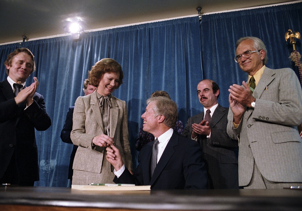 A group of people stand behind a man seated at a small writing desk. A woman stands beside him smiling as she takes a pen from him. He smiles up at her.