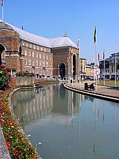 A large brick building, built in a shallow curve, with a central porch. In front of that a pool and a water fountain.