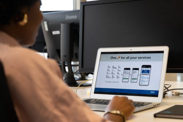 Person at a desk viewing a presentation on a laptop. Presentation screen is titled ‘One key for all your services’. Illustration on the slide shows 12 government department logos on the left with arrows pointing to three mobile screens on the right which demonstrate a mock user journey of logging-in to an online government account.