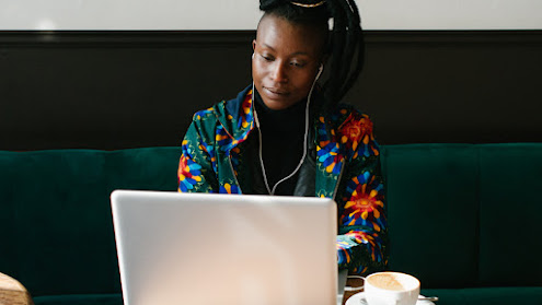 A woman of color with a colorfull jacket sit working on a laptop with earphones on and a coup of coffee at the table