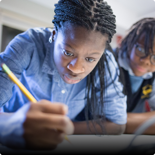 A young girl sits at a desk with a pencil in hand, working hard. Another girl in the background looks on.