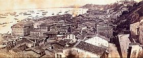 Photograph looking down a hillside overlooking the buildings of a town with a harbor beyond which is crowded with various vessels