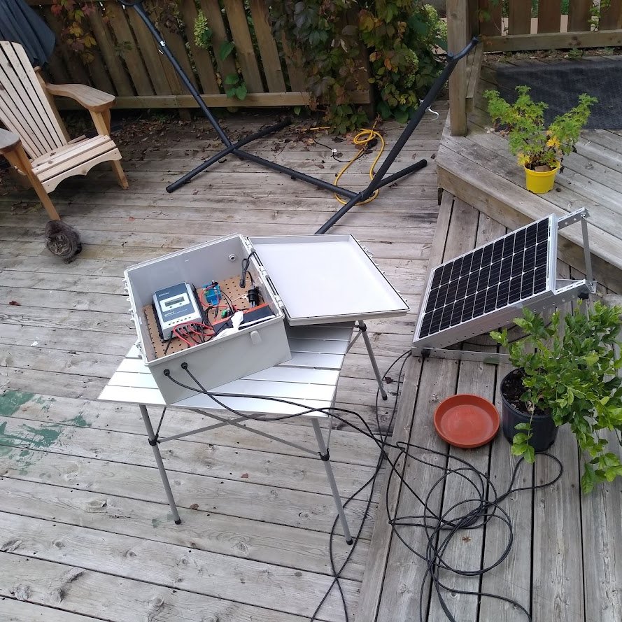 A backyard deck with a small solar panel, a grey box on a table full of electronics and a pegboard, and a small tabby cat in the background