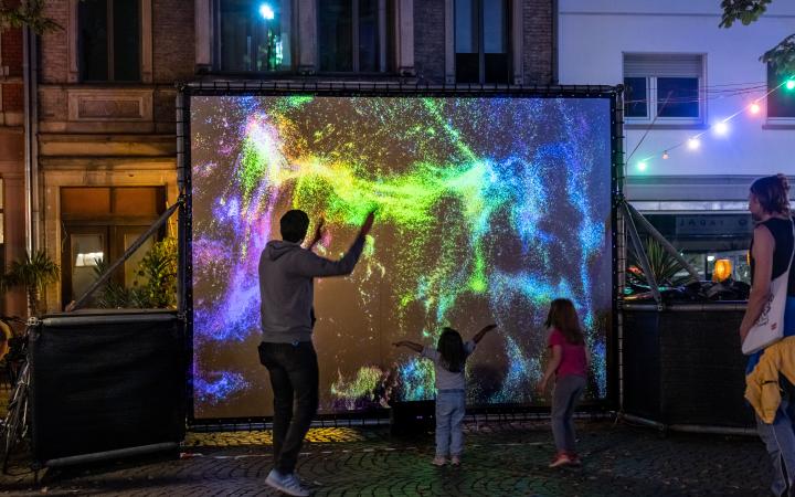 In der Fußgängerzone steht eine Leinwand mit leuchtend bunten Farbpartikeln, davor bewegen sich Erwachsene und Kinder.