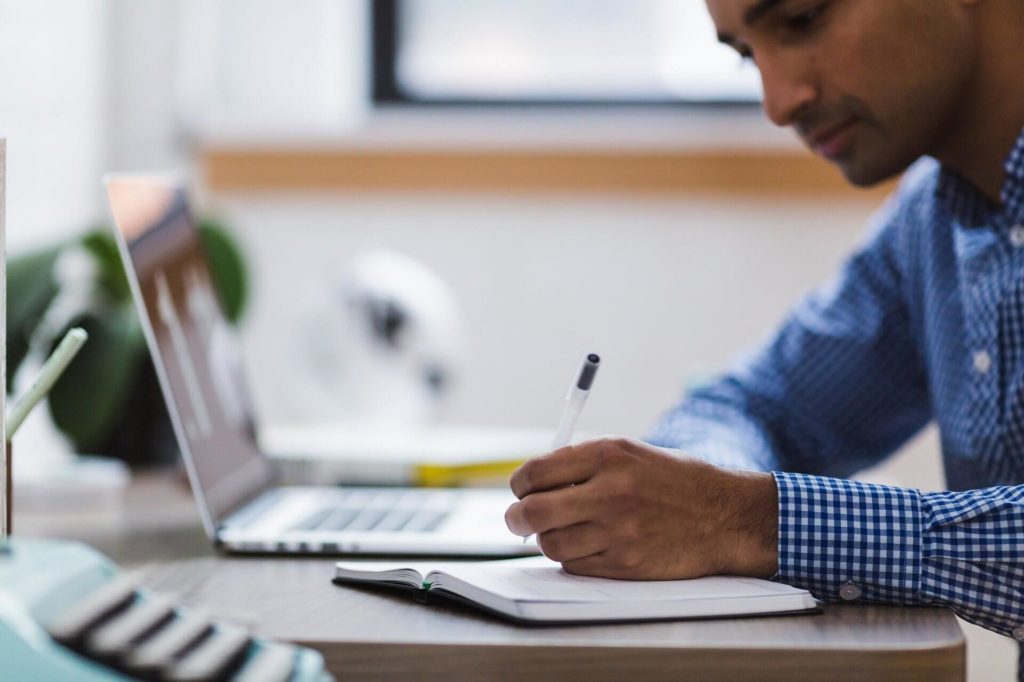 Man writing on a notepad while his laptop sits in the background.