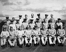 Twenty-eight Sailors in the uniform of the United States Navy pose on the deck of a World War II-era Aircraft Carrier.